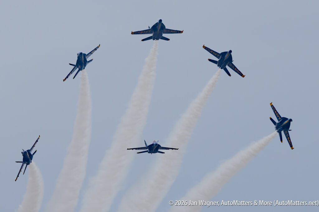 Five Blue Angels F/A-18 jets flying in formation against a clear sky, each leaving a white smoke trail.