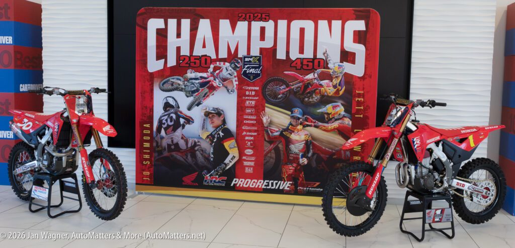 Two red Honda motocross bikes on stands flanking a large red banner commemorating 2025 SMX champions, featuring images of riders Jo Shimoda and Jett Lawrence.