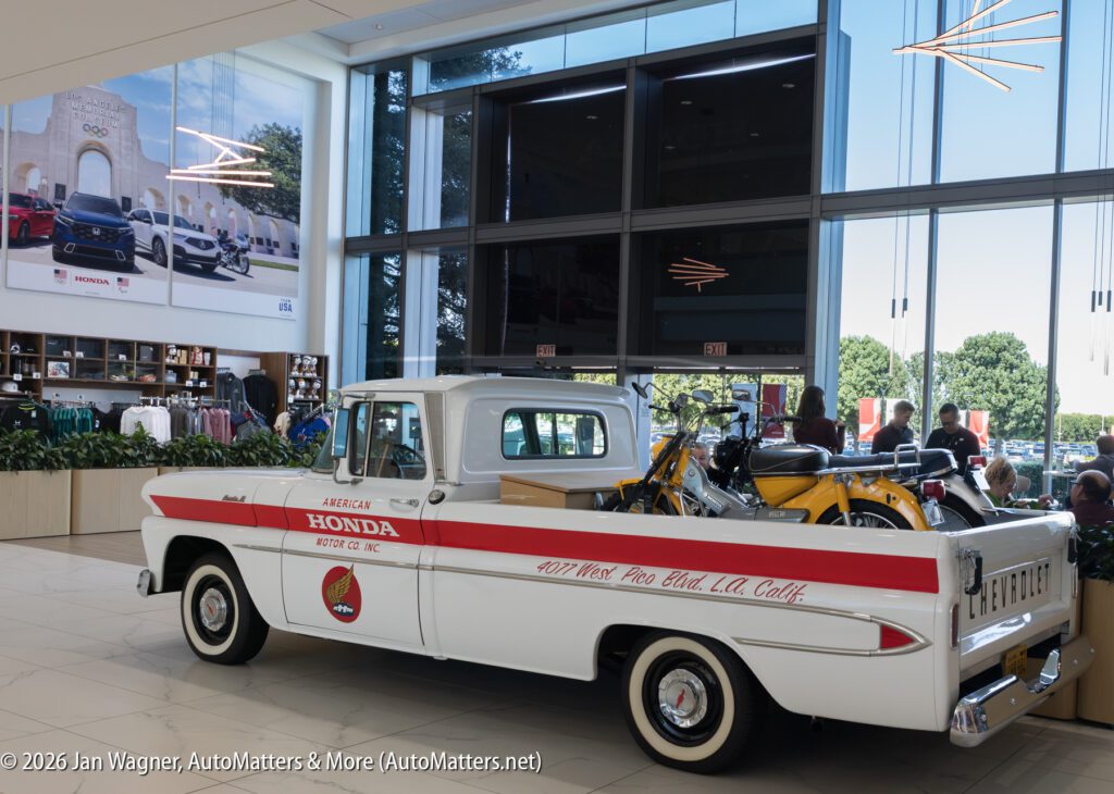 Vintage white Chevrolet pickup truck with red “American Honda Motor Co., Inc.” branding in a dealership showroom, carrying two yellow vintage Honda motorcycles in its bed.