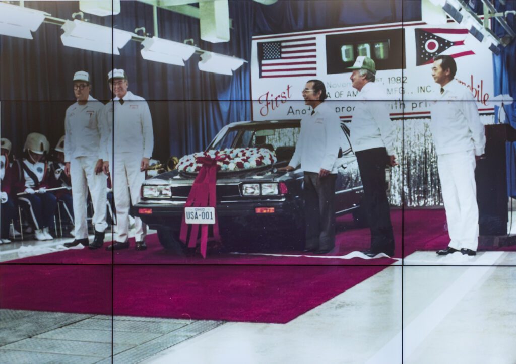 Four men in white uniforms stand beside a car decorated with a large wreath and a “USA-001” license plate, in front of a banner commemorating the first Honda of America automobile in 1982.