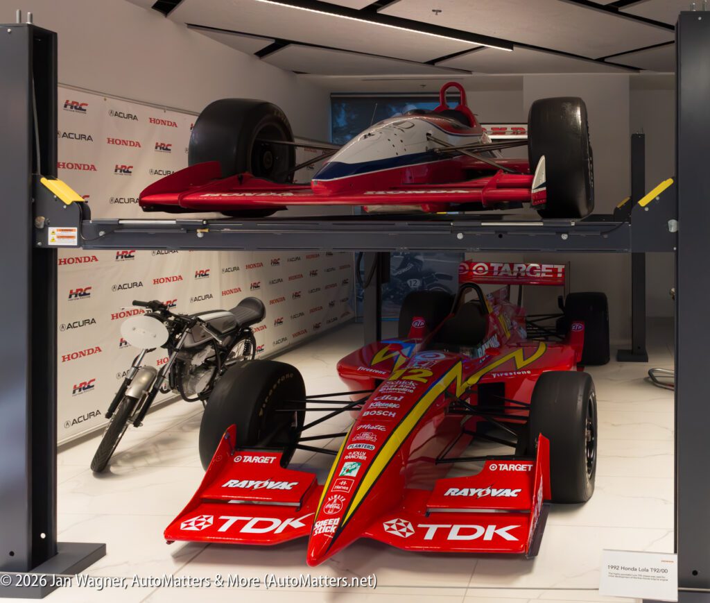 A red and white race car on a two-post lift, with a second red IndyCar and a vintage motorcycle parked on the floor beneath it.