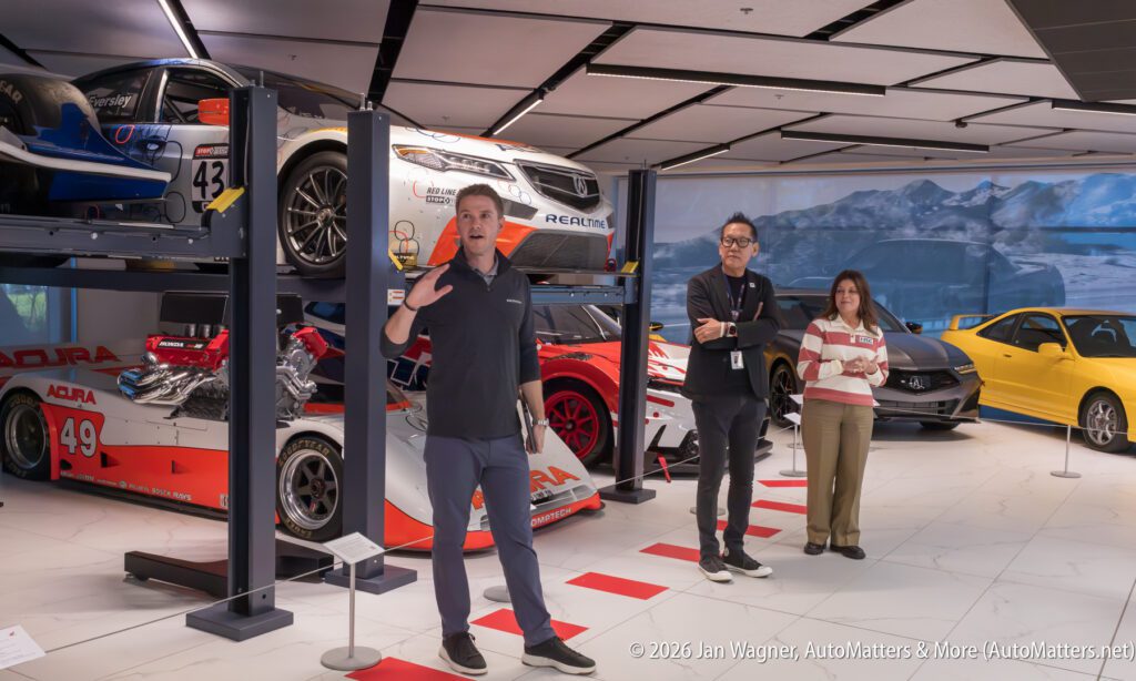 A man gestures while speaking to two people in an Acura museum gallery featuring race cars, including a stacked display.