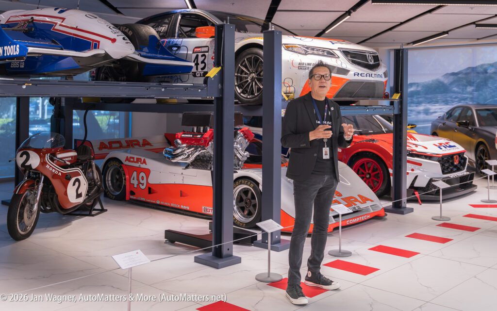 A man wearing a black blazer and glasses gestures while standing in an automotive museum displaying various Acura race cars and a vintage Honda motorcycle.