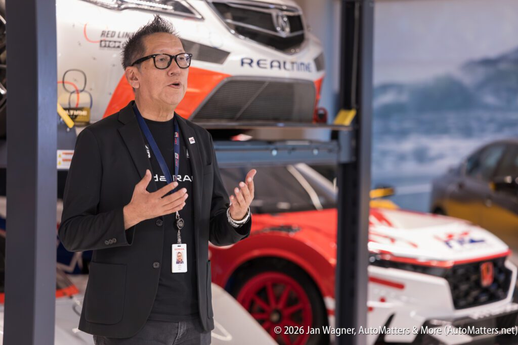 A man in a black blazer and glasses speaking with his hands, standing in a garage with race cars on a lift.