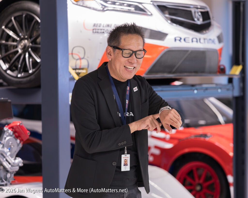 A smiling man wearing glasses and a black blazer points to his wristwatch in a garage with race cars on lifts.