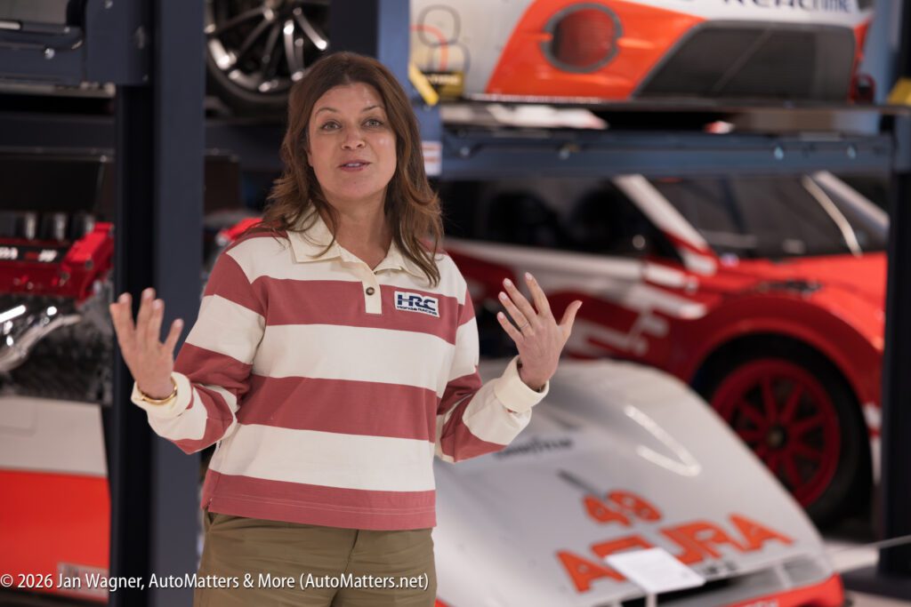 A woman in a striped shirt gestures with her hands while standing in a garage with race cars on stacked lifts.