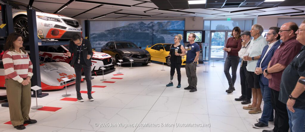 A group of people listens to a man presenting in an automotive museum featuring race cars on lifts and display platforms.