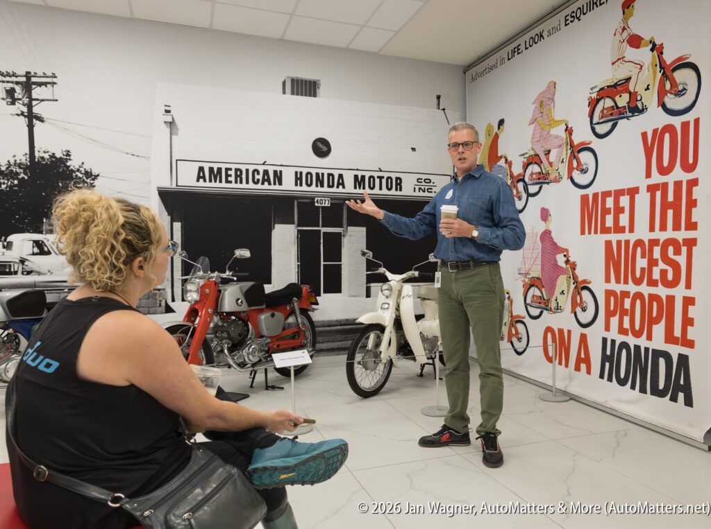 A man stands in a motorcycle showroom, gesturing toward a vintage Honda advertisement on the wall as he speaks to a seated woman.