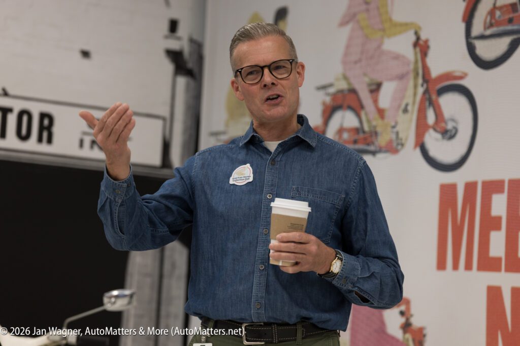 A middle-aged man in a denim shirt and glasses speaks while holding a coffee cup, standing in front of a wall with a graphic of a red motorcycle.