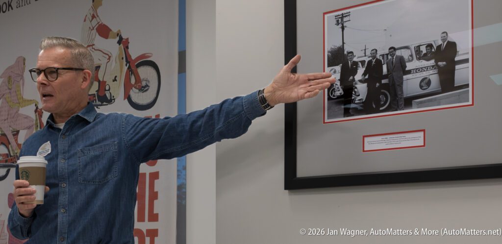 A man in a denim shirt holding a coffee cup gestures toward a framed historical photograph of American Honda Motor Co. employees.