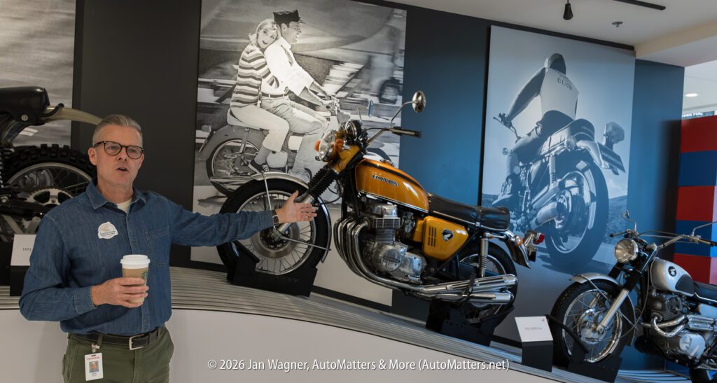 A man in a denim shirt holding a coffee cup gestures toward a vintage yellow Honda motorcycle on display in a showroom.