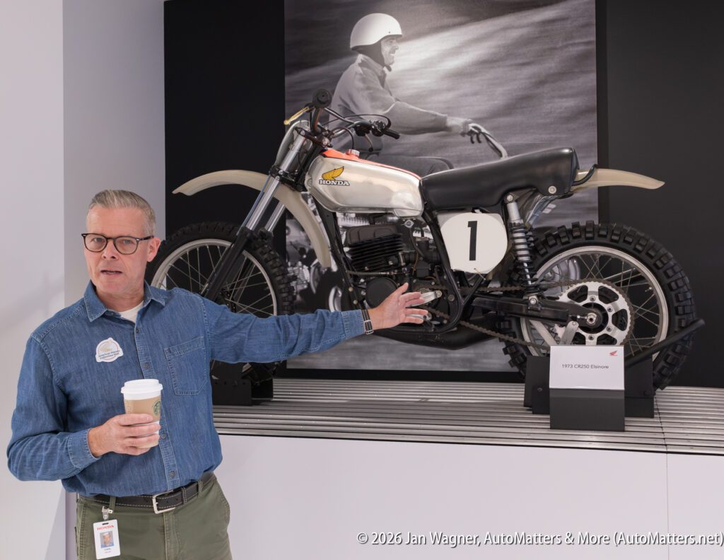 A man in a denim shirt holding a coffee cup gestures toward a vintage 1973 Honda CR250 Elsinore motocross bike displayed in front of a black-and-white photograph.