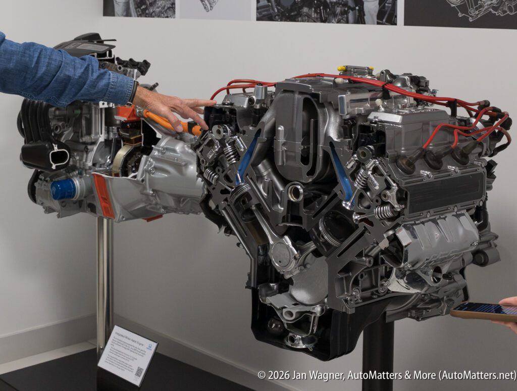 A person points to a cutaway display of a V-shaped internal combustion engine connected to an electric motor.