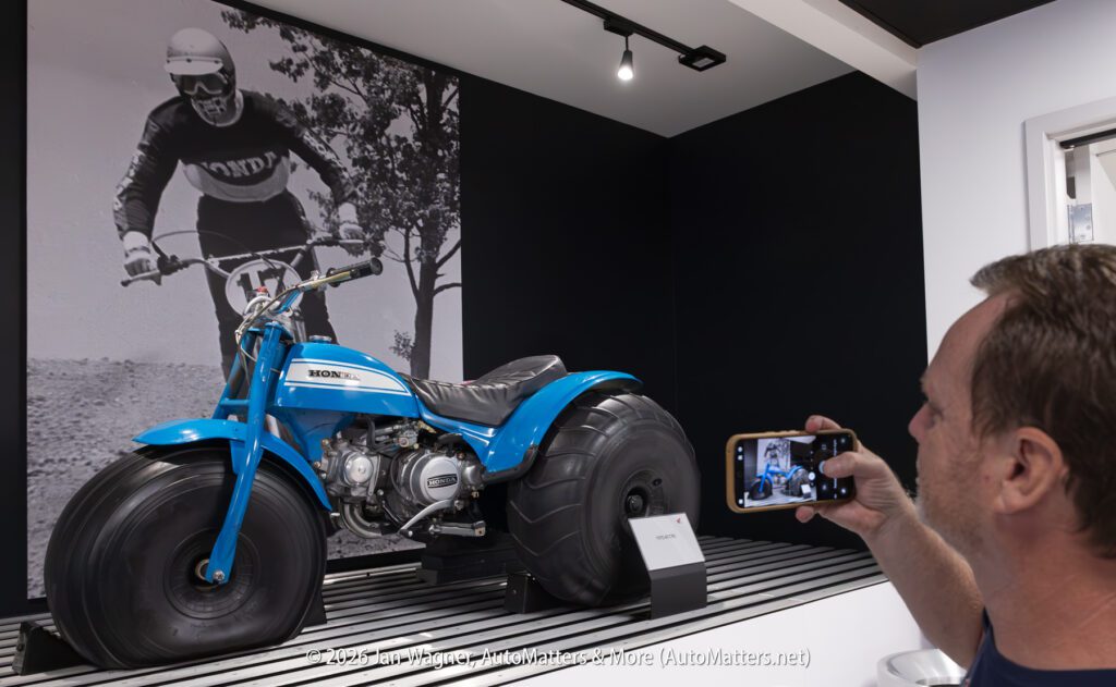 A person photographs a vintage blue Honda three-wheeled all-terrain vehicle displayed in front of a large black-and-white mural of a motorcycle rider.
