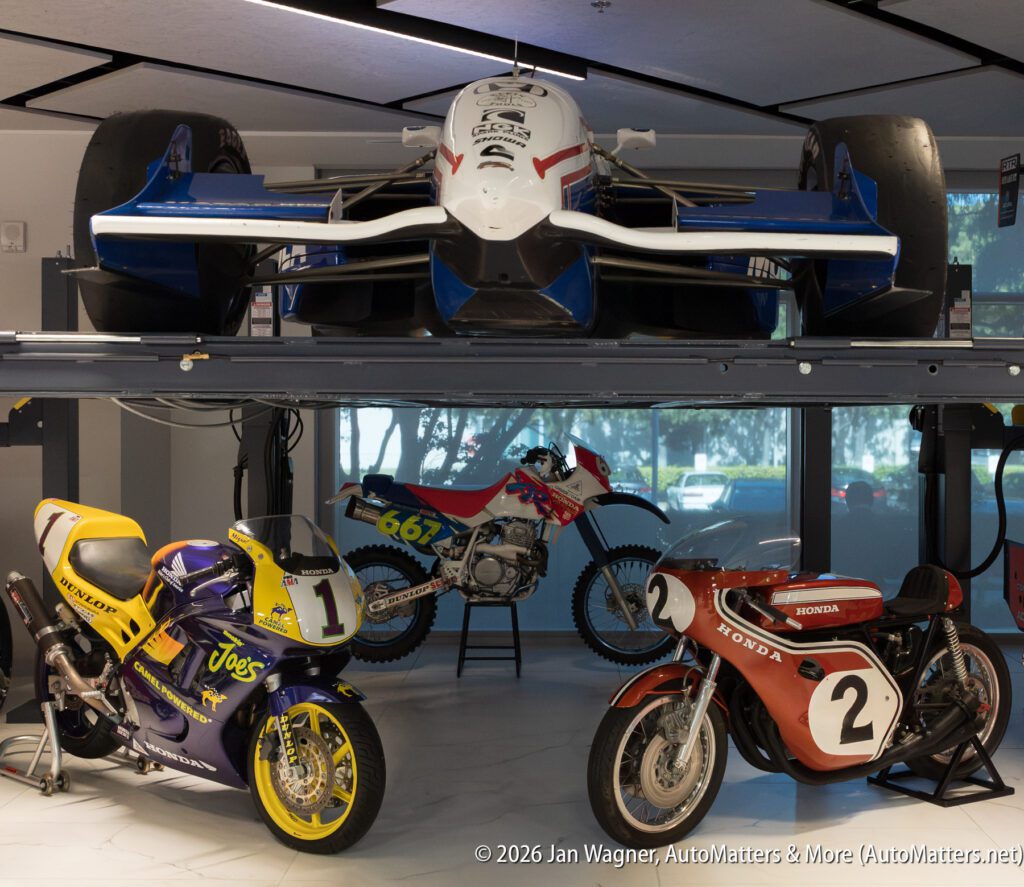 A white and blue IndyCar displayed on a lift above two Honda motorcycles: a purple and yellow racing bike and a red vintage racing bike.