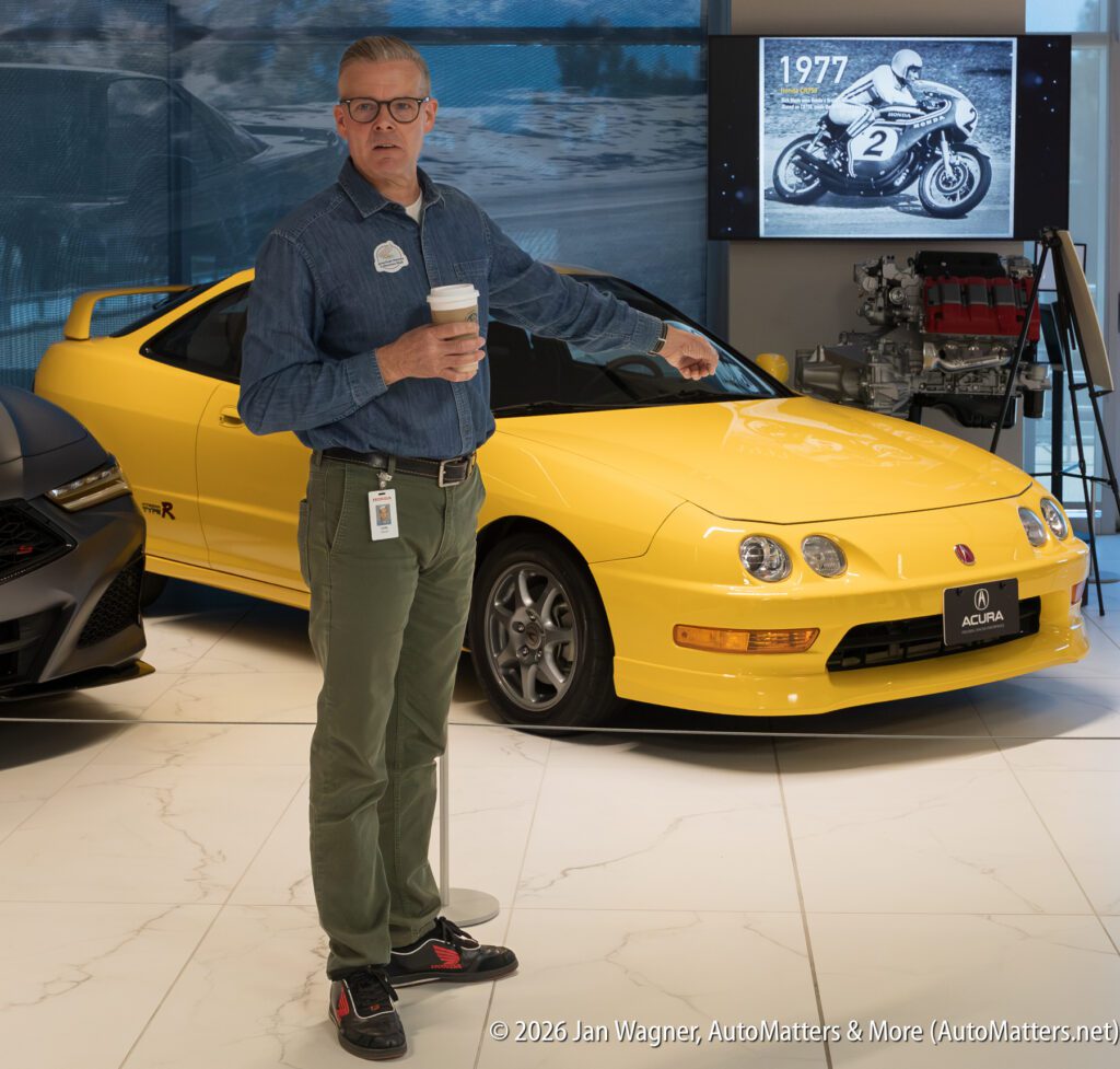 A man in a denim shirt holding a coffee cup stands in front of a yellow Acura Integra Type R in a showroom.