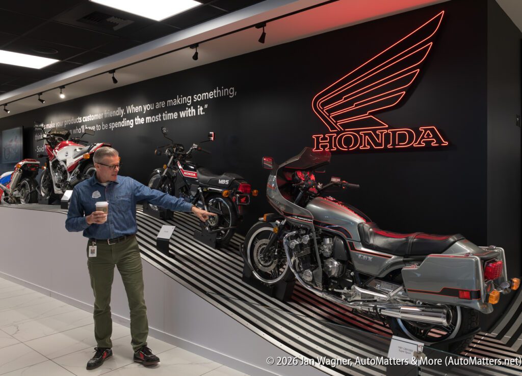 A man in a denim shirt points toward a display of vintage Honda motorcycles at a dealership, with a glowing red Honda neon sign on the wall behind him.
