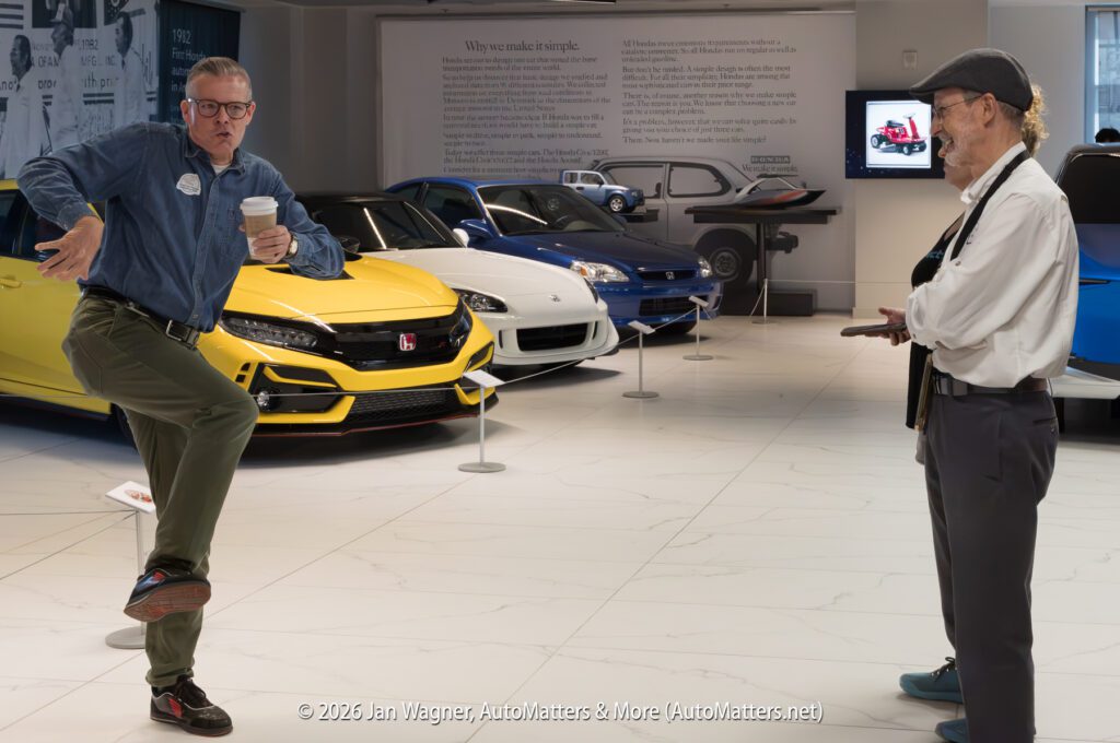 A man in a denim shirt holds a coffee cup and poses in front of a yellow Honda Civic, while another man photographs him in a car museum.