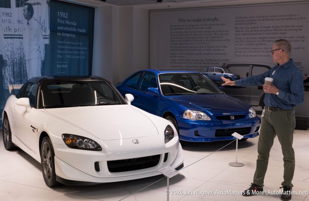 A man in a denim shirt gestures toward a white Honda S2000 and a blue Honda Civic in a museum exhibit featuring historical text panels.