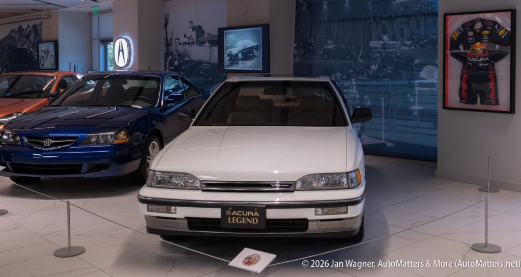 A white Acura Legend sedan on display in a showroom, with other Acura vehicles and historical wall displays visible nearby.