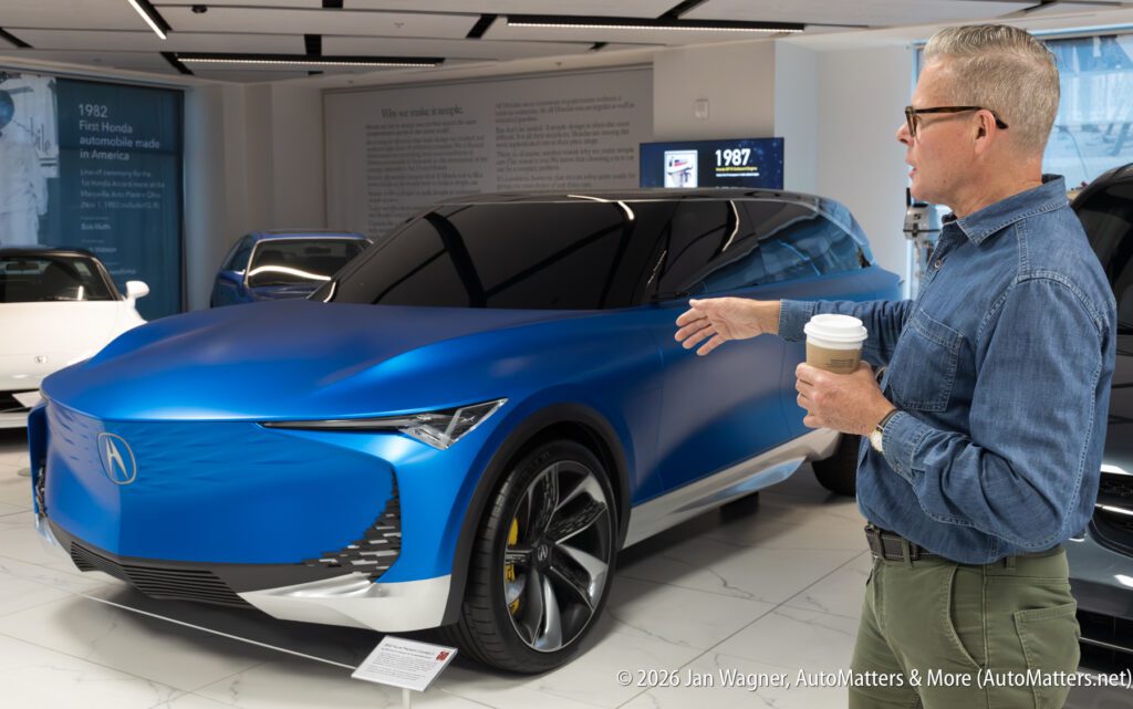 A man in a denim shirt gestures toward a blue Acura concept SUV on display in a showroom.
