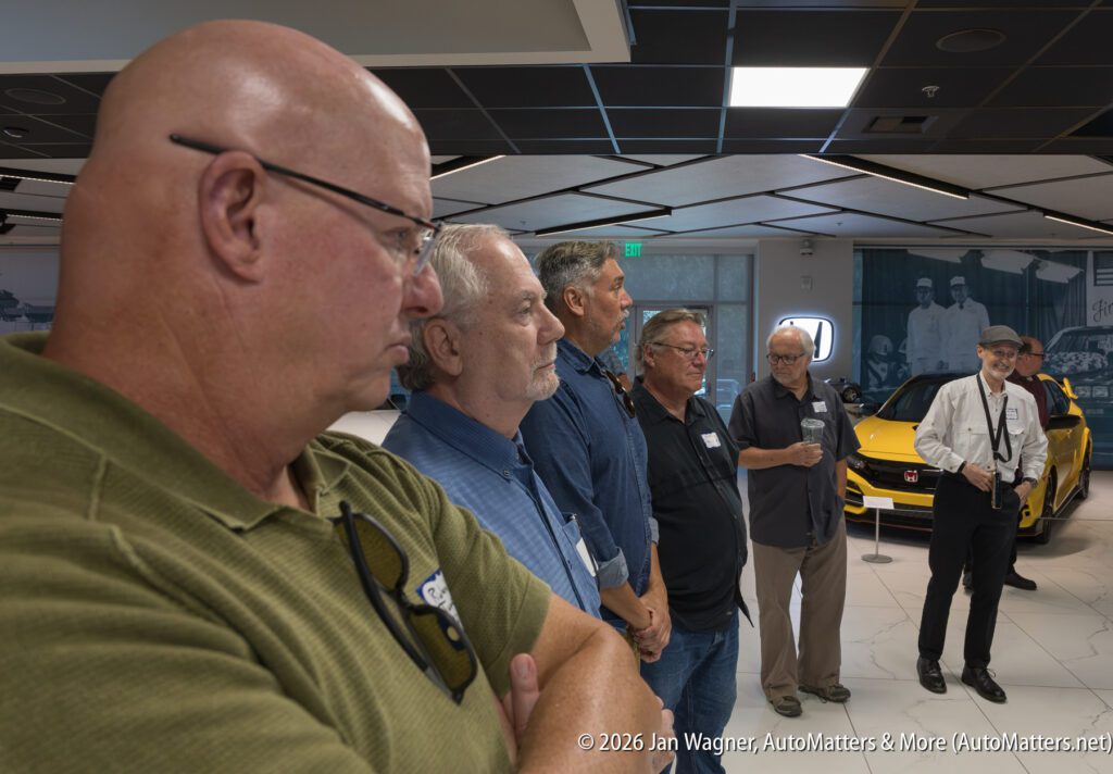 Five men stand in a car dealership showroom looking toward a yellow Honda Civic Type R.