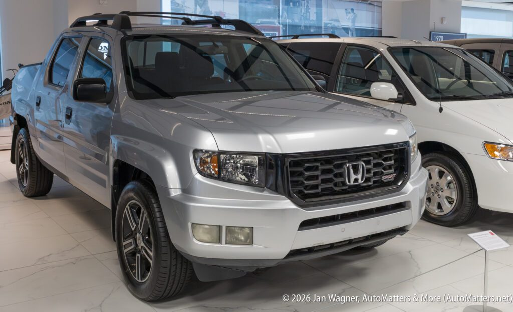 Silver Honda Ridgeline truck parked in a showroom next to a white minivan.