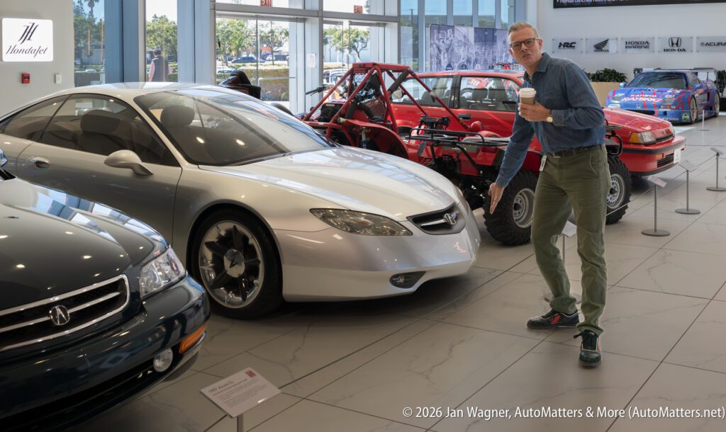 A man in a denim shirt and green pants gestures toward a silver Acura concept car in a dealership showroom.