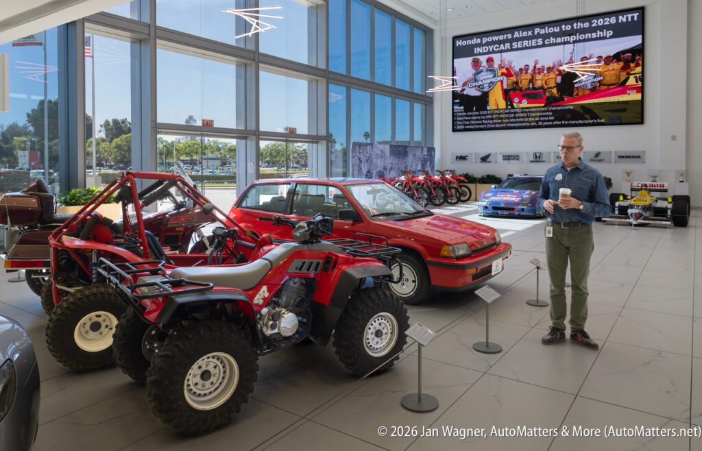 A man holding a coffee cup stands in a museum gallery featuring several red Honda vehicles, including an ATV, a hatchback, and motorcycles, with a digital display about IndyCar racing on the wall.