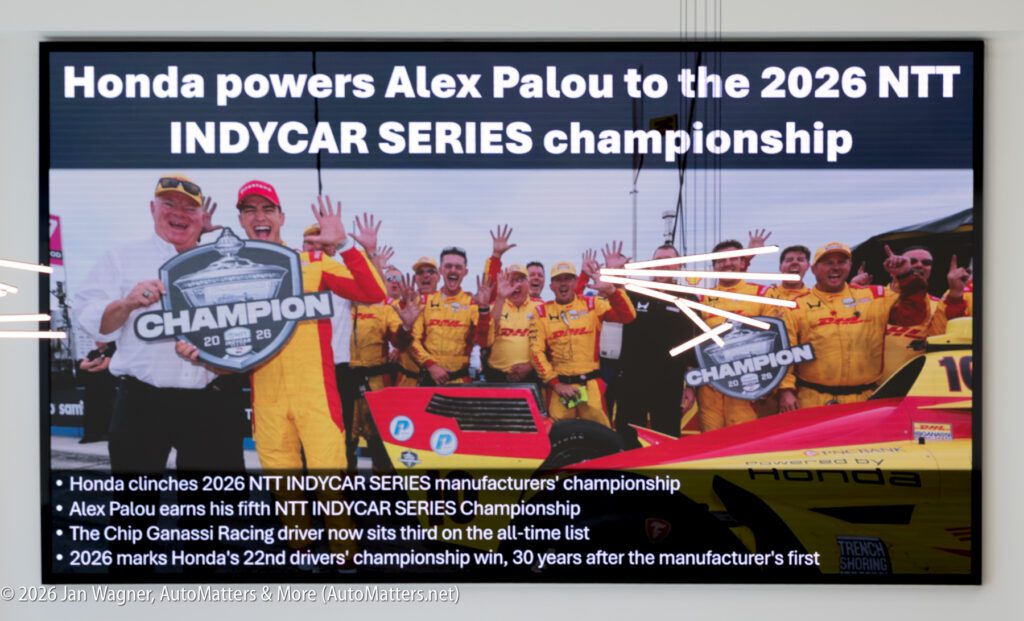 A digital display showing race car driver Alex Palou and his team celebrating his 2026 NTT INDYCAR SERIES championship win, with bulleted text highlighting the team’s achievements.