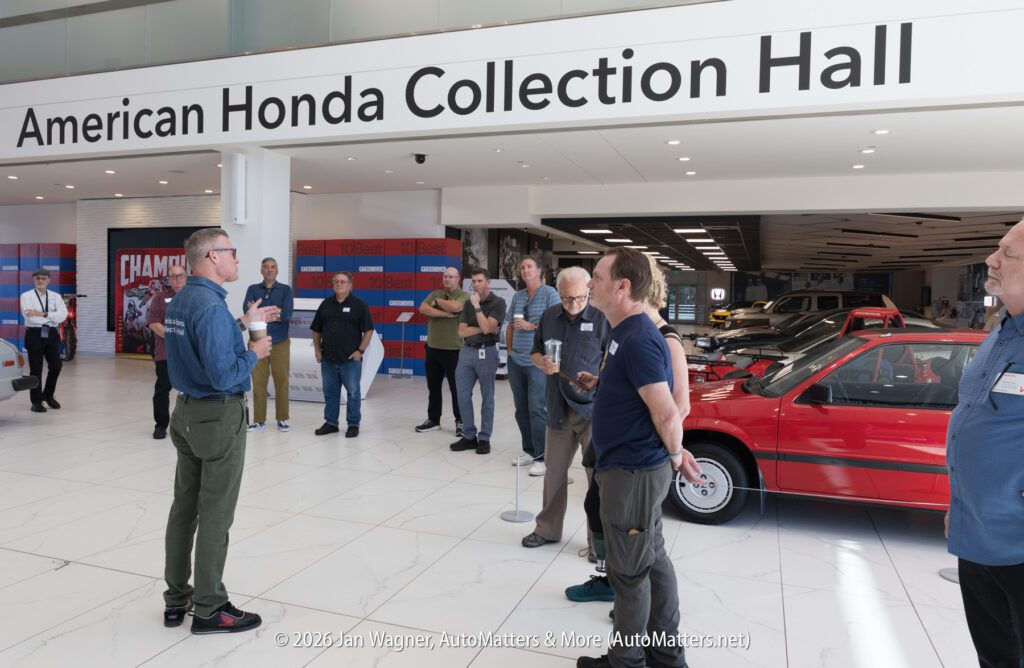 A group of people standing in the American Honda Collection Hall, listening to a man speaking near a red vintage Honda car.
