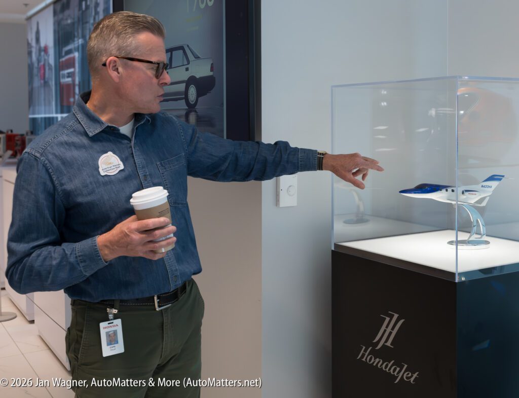A man in a denim shirt holding a coffee cup points to a HondaJet model airplane inside a glass display case.