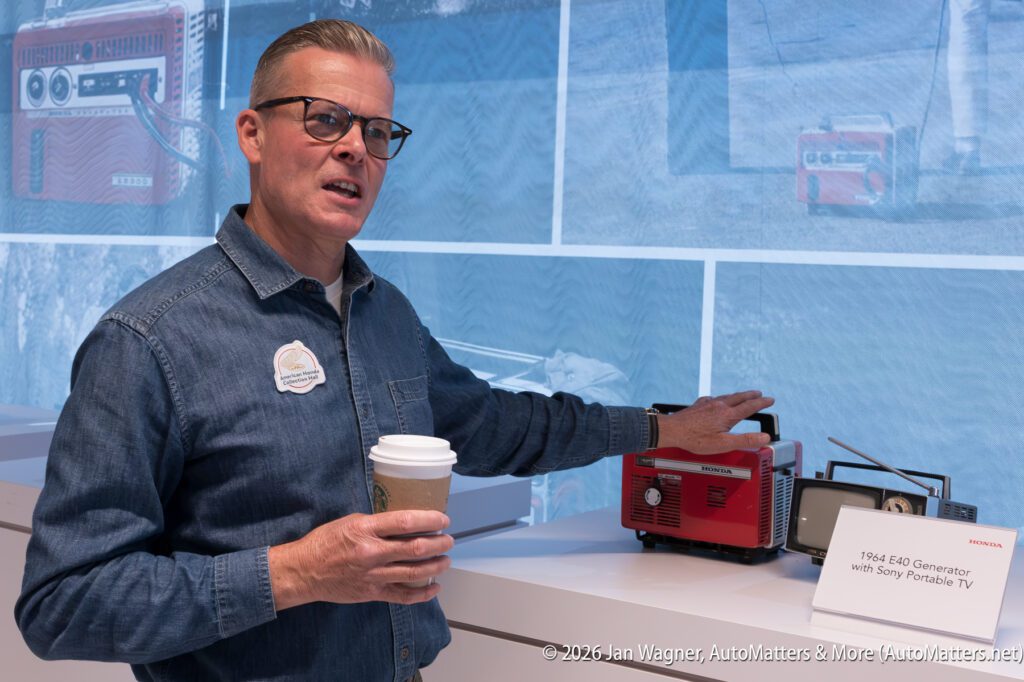 A man in a denim shirt stands next to a small red 1964 Honda E40 generator and a portable television, holding a coffee cup.