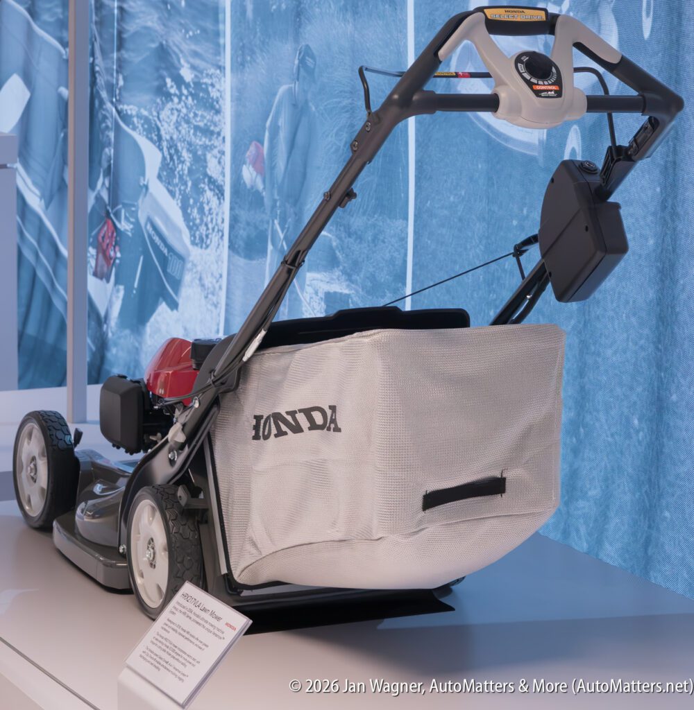 A Honda lawn mower with a grass collection bag, displayed on a platform in front of a blue wall graphic.