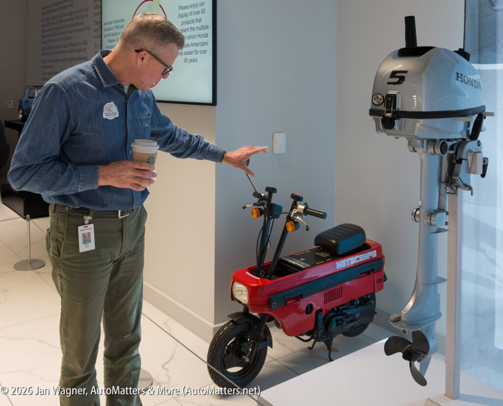 A man in a denim shirt holding a coffee cup points to a red Honda Motocompo scooter next to a Honda outboard motor.