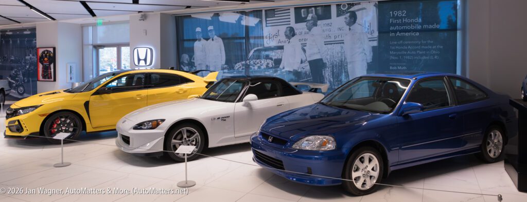Three Honda cars parked in a showroom, including a yellow Civic Type R, a white S2000, and a blue Civic Si coupe, in front of a wall featuring historical photos and text about Honda history.