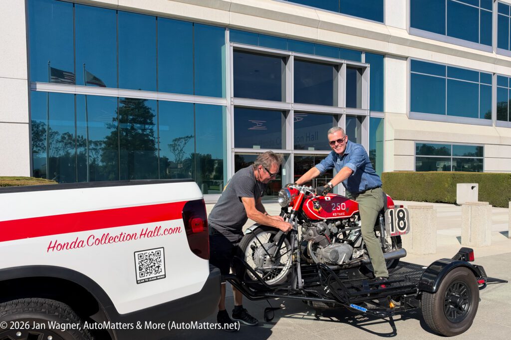 A man sits on a vintage red Honda 250 motorcycle on a trailer while another man secures the front wheel, parked in front of the Honda Collection Hall.